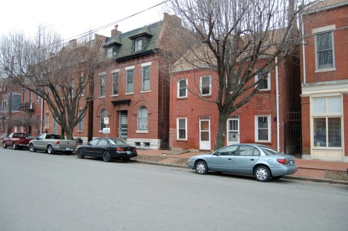 A row of brick houses with bare trees lining the street, parked cars along the curb on a cloudy day.