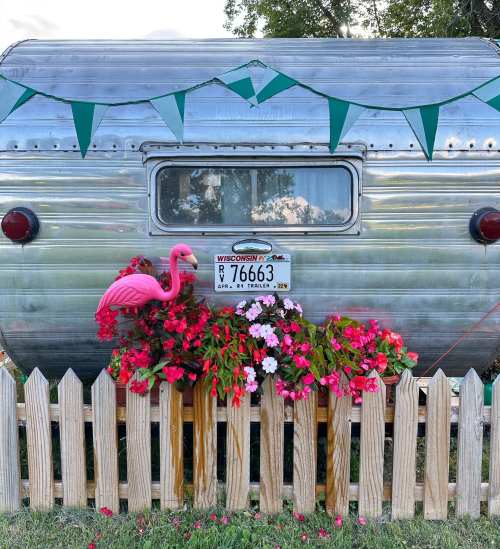 A vintage silver trailer with a pink flamingo and colorful flowers in front, decorated with green bunting.