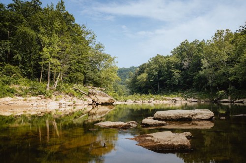 A serene river scene with smooth rocks, lush green trees, and a clear blue sky reflecting on the water's surface.
