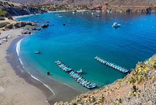 A serene beach with clear blue water, lined with boats and rocky cliffs in the background.
