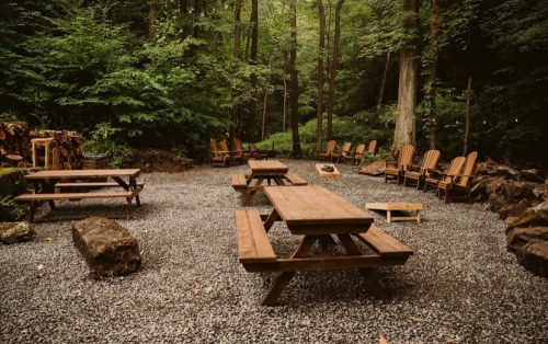 A serene outdoor space with wooden picnic tables and chairs surrounded by lush greenery and gravel pathways.