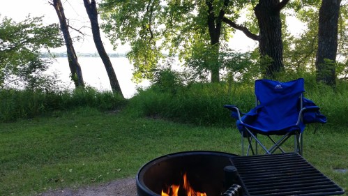 A campfire burns in a fire pit beside a lake, with a blue camping chair and trees in the background.