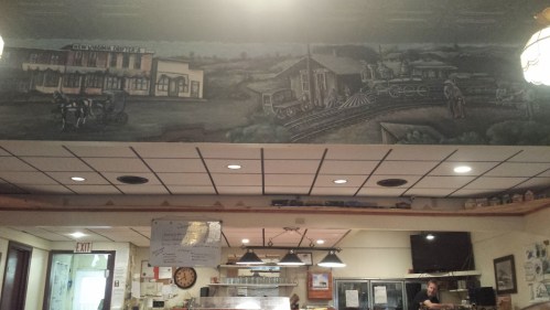 Mural depicting a historic scene with a train and horse-drawn carriage, above a cozy diner interior.