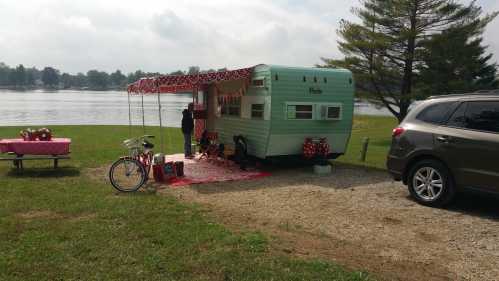 A vintage teal camper by a lake, decorated with red and white accents, alongside a picnic setup and a bicycle.