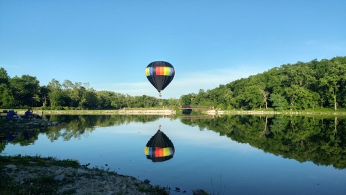 A colorful hot air balloon floats above a calm lake, reflecting on the water, surrounded by lush greenery.