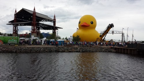 A large yellow rubber duck floats near a crowd by the water, with a stage and structures in the background.