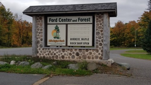 Sign for Ford Center and Forest, featuring a logo and information about Birdeye Maple and a rock shop. Autumn trees in background.