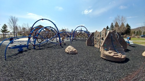 A playground featuring climbing structures, slides, and a rock wall, surrounded by a rubber surface and trees in the background.