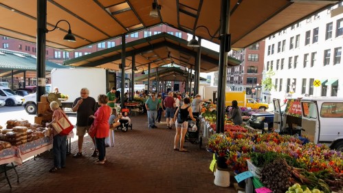 A bustling farmers market with vendors, shoppers, and colorful flower displays under a large canopy.