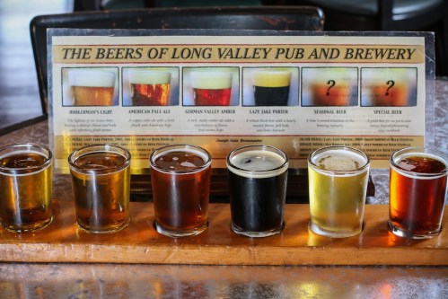 A wooden flight board with six small glasses of different beers in front of a menu displaying various beer options.