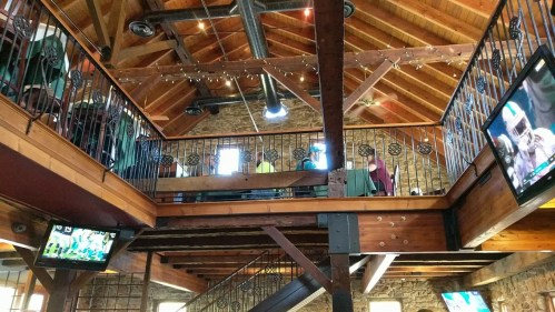 Interior of a rustic venue with wooden beams, two levels, and TVs displaying sports, creating a lively atmosphere.