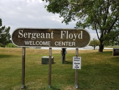 Sign for Sergeant Floyd Welcome Center, with hours displayed: Open daily 10 AM to 4 PM, surrounded by greenery.