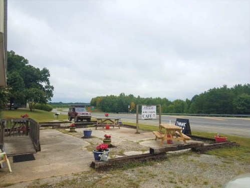 A roadside café with picnic tables, flower pots, and a vintage truck parked nearby, set against a cloudy sky.