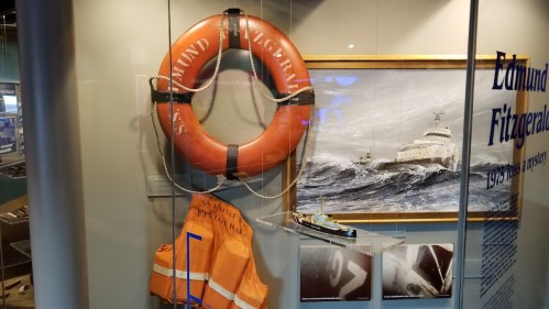 Lifebuoy and orange life jacket displayed alongside a painting of the Edmund Fitzgerald in a museum exhibit.