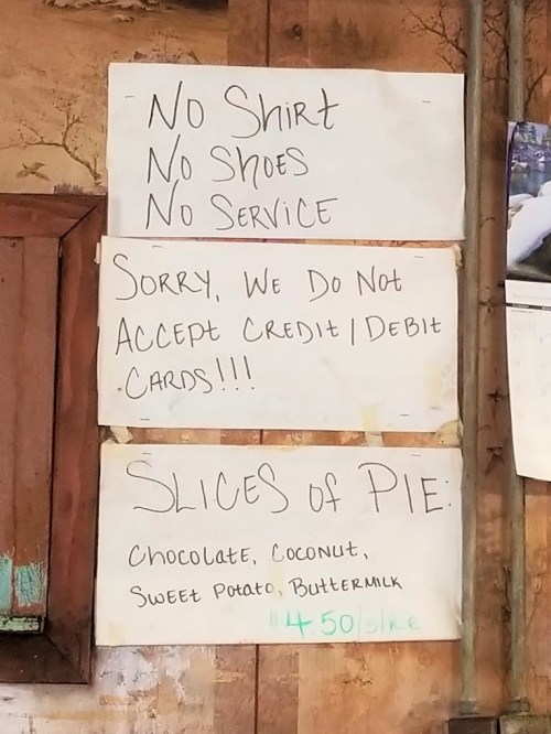 Sign on a wall with rules: "No Shirt, No Shoes, No Service" and a menu for pie slices with prices.