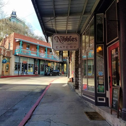 Sign for "Nibbles Eatery" on a street with colorful buildings and shops in the background.
