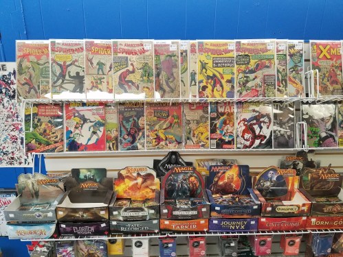A display of comic books featuring Spider-Man and X-Men, with various trading card games below.