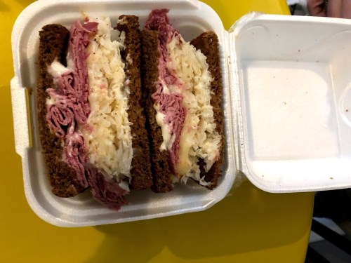 A sandwich with layers of corned beef, sauerkraut, and cheese on dark rye bread, served in a takeout container.