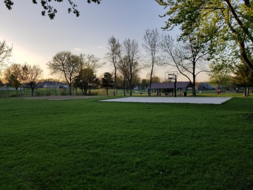 A grassy park with trees, a basketball court, and a pavilion under a clear sky during sunset.