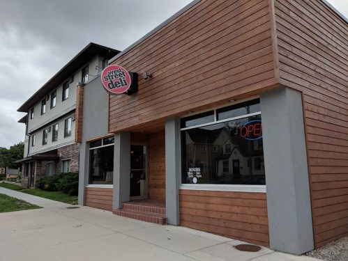 A modern deli with a wooden facade and an "OPEN" sign, located next to a multi-story building.