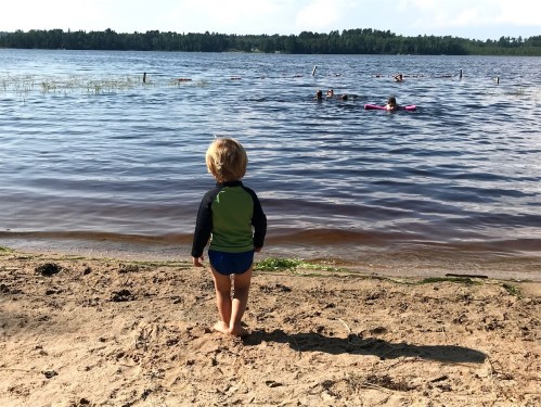 A child stands on the sandy shore, watching people swim in a lake on a sunny day.