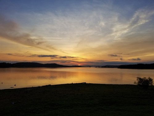 A serene sunset over a calm lake, with soft clouds and silhouettes of distant hills. Ducks swim in the foreground.