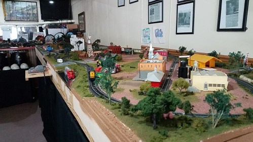 A detailed model train layout featuring buildings, trees, and tracks in a room with framed photos on the walls.