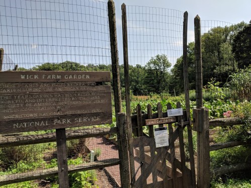 A wooden sign marks the entrance to Wick Farm Garden, surrounded by a fenced garden area with greenery in the background.