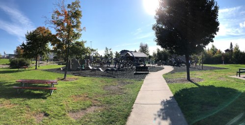 A sunny park scene featuring a playground, walking path, and picnic tables surrounded by trees and green grass.