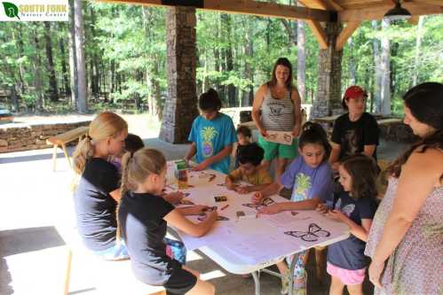 A group of children and adults gather around a table, engaged in arts and crafts in a wooded outdoor setting.