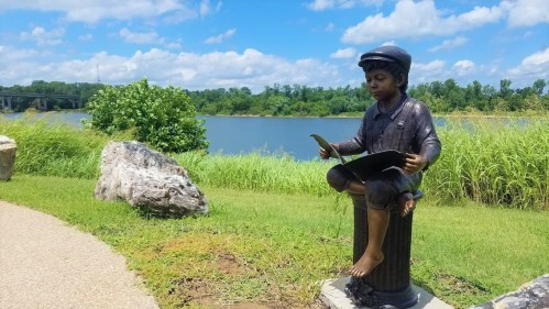 A bronze statue of a boy reading sits on a pedestal by a lake, surrounded by greenery and blue skies.