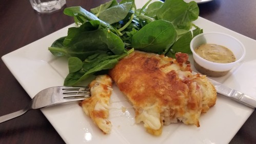A plate with a piece of golden-brown baked fish, fresh spinach, and a small cup of dressing on the side.