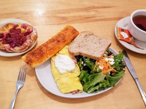 A plate with an omelette, salad, bread, and a pastry, alongside a cup of tea with sugar packets.