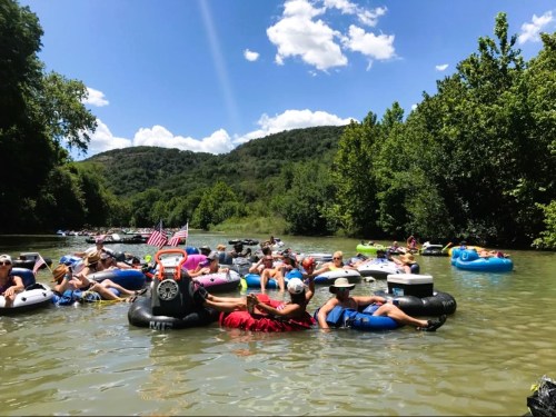 A group of people floating on inner tubes in a river, surrounded by trees and hills under a blue sky with clouds.