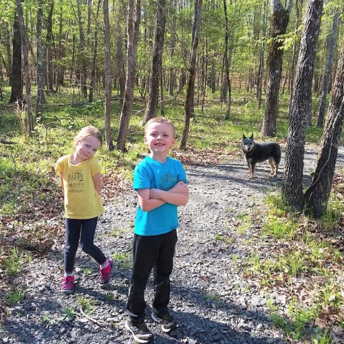 Two children stand on a forest path, smiling, with a dog nearby among trees and greenery.