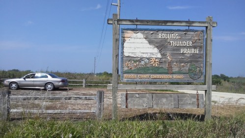 Sign for Rolling Thunder Prairie, with a car parked nearby and a clear blue sky in the background.