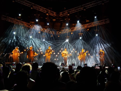 A band performs on stage with bright lights and a cheering crowd in front, creating an energetic concert atmosphere.