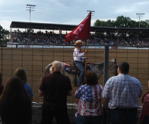 A rider on a horse holds a flag in a rodeo arena, with a crowd watching from behind a fence.