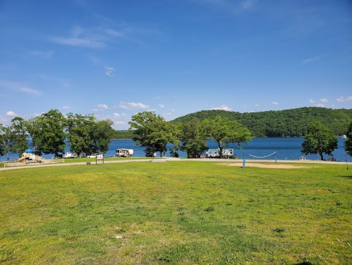 A serene lakeside view with green grass, trees, and RVs parked along the water under a clear blue sky.