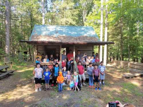 A large group of children and adults gathered outside a rustic cabin in a wooded area, smiling for the camera.