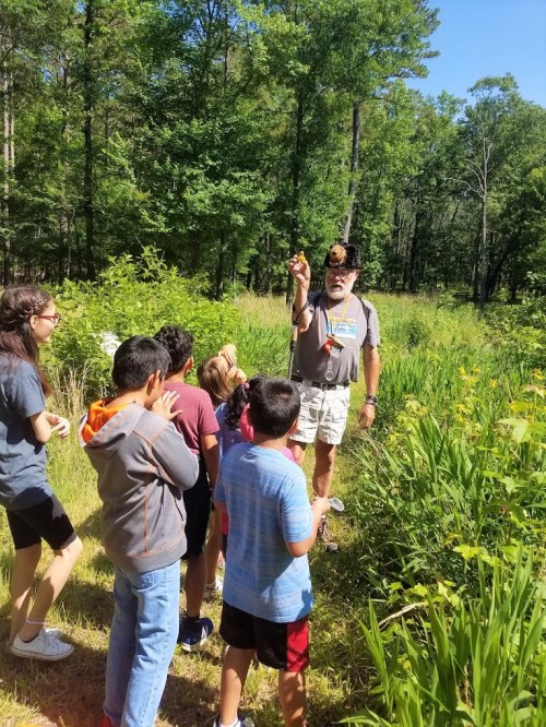 A man in a hat shows a flower to a group of children in a lush, green outdoor setting.