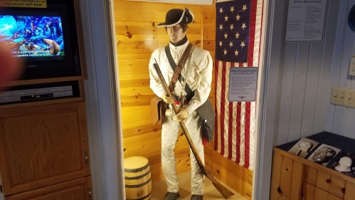 A life-sized mannequin of a Revolutionary War soldier stands beside an American flag and a wooden barrel.