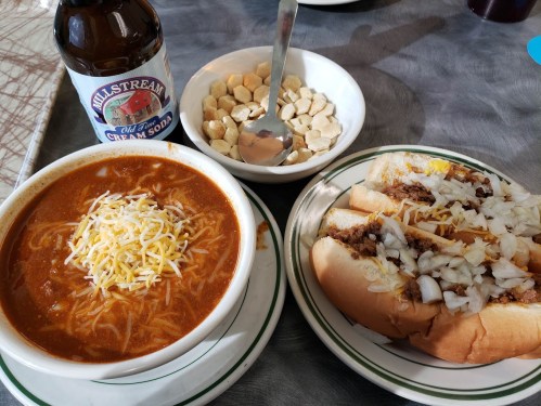 A plate with chili cheese dogs topped with onions, a bowl of crackers, and a bottle of cream soda.