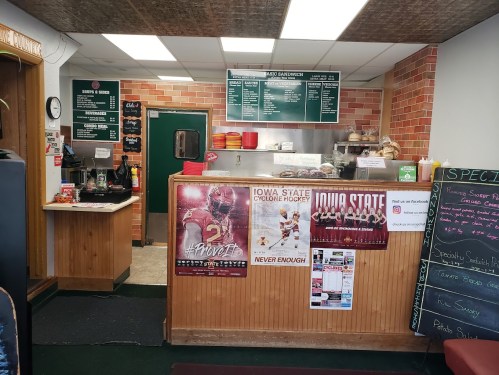 Interior of a sandwich shop featuring a menu board, counter, and posters for Iowa State sports on the walls.