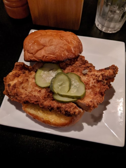 A fried chicken sandwich with pickles on a toasted bun, served on a white plate.