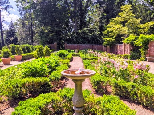 A serene garden with lush greenery, flower beds, and a stone fountain in the foreground under a clear blue sky.