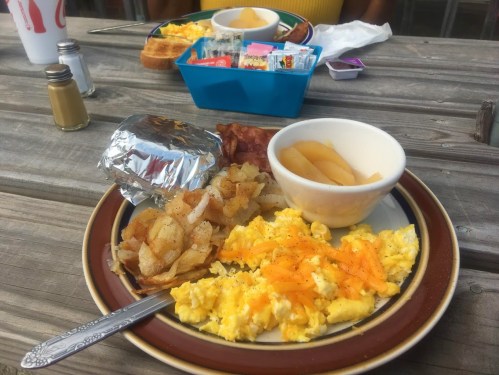 A plate of scrambled eggs with cheese, hash browns, bacon, a biscuit, and a bowl of canned peaches on a wooden table.