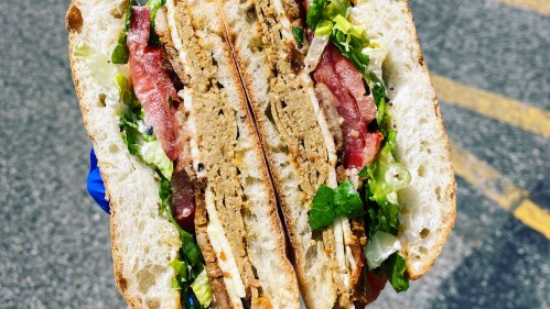 A close-up of a sandwich filled with layers of lettuce, tomato, and a textured protein patty, held in a hand.