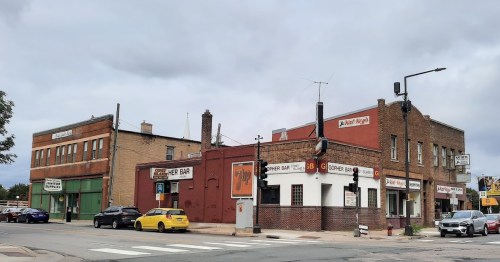 A street view of a corner bar and nearby buildings under a cloudy sky, with parked cars along the road.