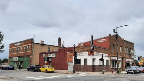 A street view of a corner bar and nearby buildings under a cloudy sky, with cars parked along the road.